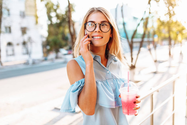 Young beautiful girl student, wearing glasses, talking on the phone in a good mood, and holding a cocktail - Foto, immagini