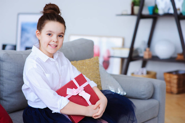 счастливая девочка с подарком на день рождения или Рождество позирует дома в современном скандинавском интерьере, готовит или разворачивает праздничный подарок. - Фото, изображение