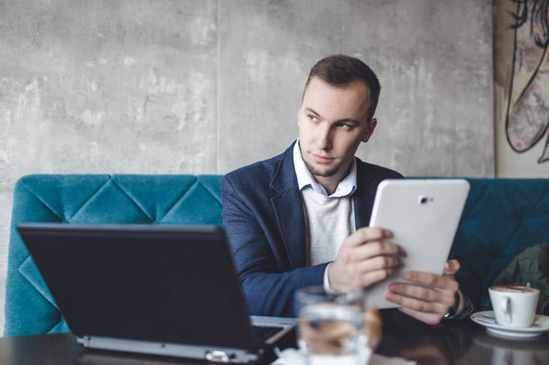 Νέοι, σύγχρονη και θετική επιχείρηση άνθρωπος κάθεται στο καφενείο και να κάνει τη δουλειά του στο σημειωματάριο, κινητό τηλέφωνο και tablet. - Φωτογραφία, εικόνα