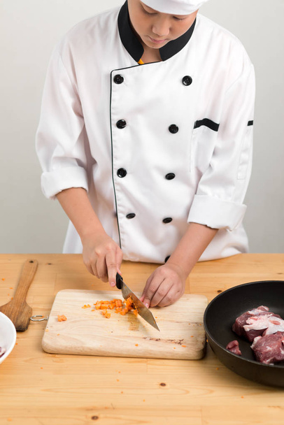Asian preteen boy in chef uniform chopping carrot on table, preparing food. - Photo, image