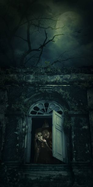 Halloween witch holding a skull standing in ancient window building over dead tree, birds, full moon and spooky cloudy sky, Halloween mystery concept - Photo, Image