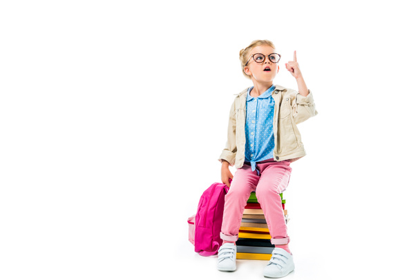niño escolar emocionado apuntando hacia arriba y teniendo idea mientras está sentado en un montón de libros con mochila aislada en blanco, estudiando concepto
  - Foto, imagen