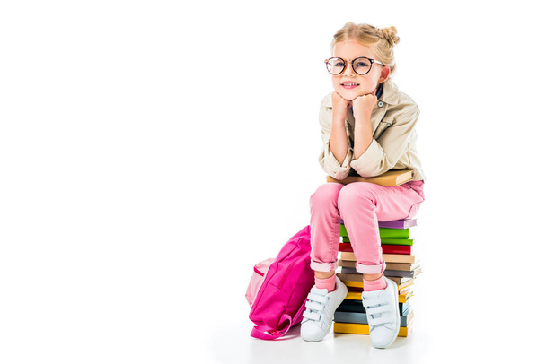 adorable smiling schoolgirl sitting on pile of books isolated on white  - Foto, afbeelding