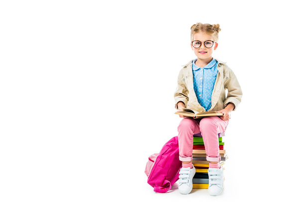 adorable pupil reading while sitting on pile of books with pink backpack isolated on white - Фото, зображення