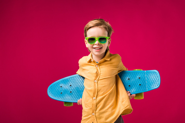 stylish cheerful boy in sunglasses posing with penny board isolated on red - Photo, Image