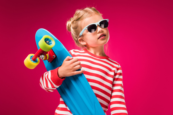 stylish kid in sunglasses posing with skateboard isolated on red - Foto, imagen