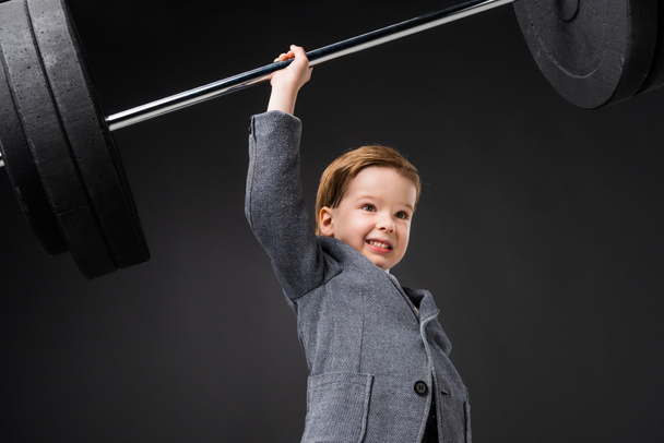 strong cheerful boy in suit lifting barbell isolated on grey - Fotó, kép