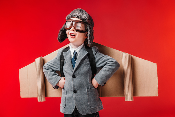 збуджений хлопчик у костюмі з паперовими плоскими крилами та окулярами ізольовані на червоному
 - Фото, зображення