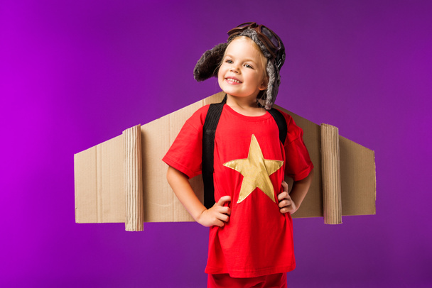 little cheerful pilot with cardboard plane wings and goggles isolated on purple - Photo, Image
