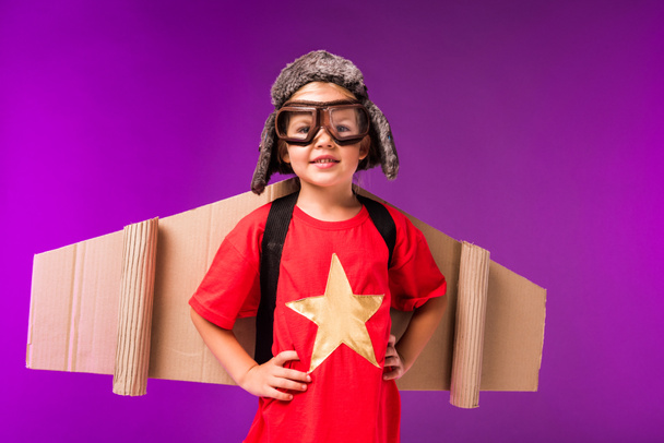 boldog gyermek papír repülőgép szárnyak, kalap és elszigetelt lila védőszemüvegek - Fotó, kép