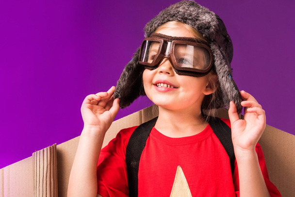 uśmiechnięte dziecko ze skrzydeł samolotu papieru i okulary ochronne na fioletowym tle - Zdjęcie, obraz