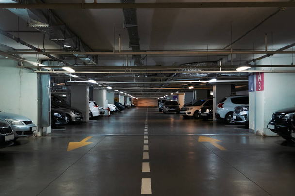 Illuminated underground car parking interior under modern mall with lots of vehicles and arrows on floor - Photo, Image