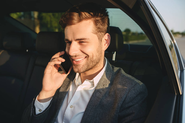 Portrait of successful man 30s in classic suit talking on smartphone while back sitting in business class car - Photo, Image