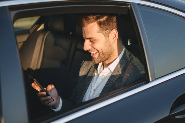 Foto de hombre feliz en traje clásico sosteniendo y utilizando el teléfono móvil mientras está de vuelta sentado en el coche de clase empresarial
 - Foto, Imagen