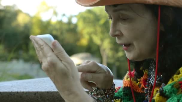 Volwassen stijlvolle vrouw in een stijlvolle cowboyhoed en een multi gekleurde poncho gebruikt haar smartphone in een park. Concept van actieve recreatie voor middelbare leeftijd en oudere mensen. - Video
