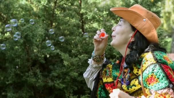 Mujer con estilo adulto en un sombrero de vaquero y un poncho multicolor sopla burbujas en el parque al aire libre. Concepto de ocio activo para personas de mediana edad y mayores
. - Metraje, vídeo