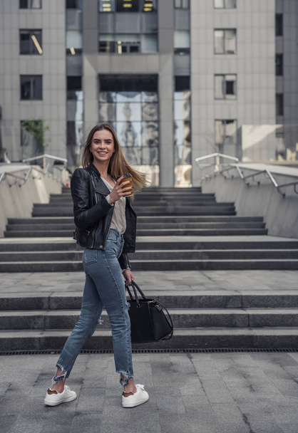 Beautiful young girl in casual clothes is holding cup of coffee, looking at camera and smiling while walking in the city center, full-length - Photo, Image