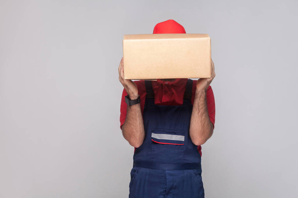Portrait of young logistic delivery man in blue uniform and red t-shirt standing and covering face with cardboard box on grey background - Photo, image