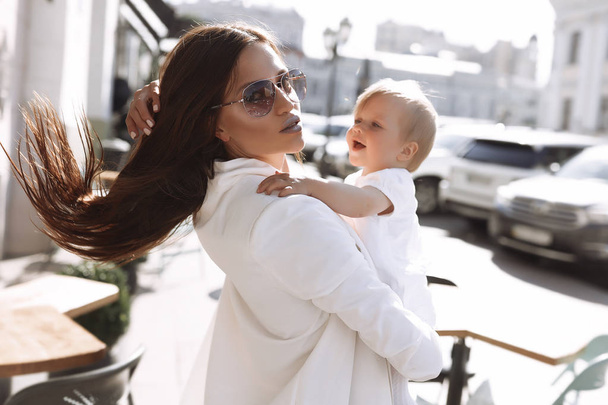 Beautiful woman and her little girl, hold her, giving tender and love, funny kid, stylish hair, hugging with mother, red cheeks, white clothes, sun glasses, brunette, sunny day, fashion mom - Foto, Imagen