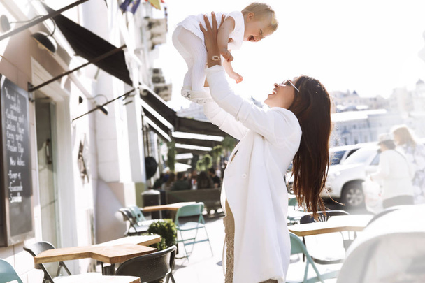 Mother and daughter are playing, put the baby up, laughing and smiling family, tender and love, funny kid, stylish hair, red cheeks, white clothes,  brunette, sunny day, fashion mom and kid, baby girl - Foto, Imagen