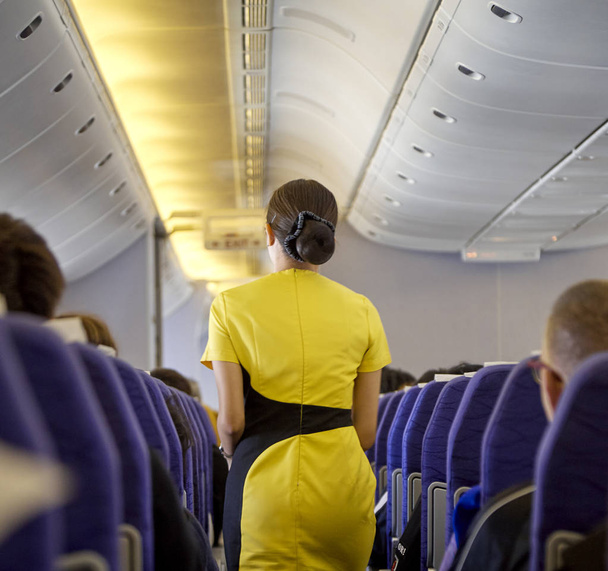 Interior del avión comercial con el vuelo attandant que sirve a pasajeros en asientos durante vuelo. Azafata en uniforme amarillo oscuro caminando por el pasillo. Composición horizontal
. - Foto, imagen