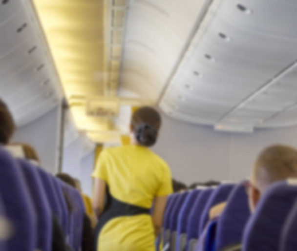 Das unscharfe Innere eines Verkehrsflugzeugs mit einem Flugbegleiter, der die Passagiere während des Fluges auf den Sitzen bedient. Stewardess in dunkelgelber Uniform durch den Gang. horizontale Zusammensetzung. - Foto, Bild