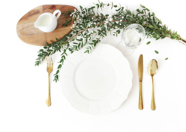 Festive wedding, birthday table setting with golden cutlery, eucalyptus parvifolia, glass of wine and milk pitcher. Bempty porcelain plate mockup. Rustic restaurant menu concept. Flat lay, top view - Foto, imagen