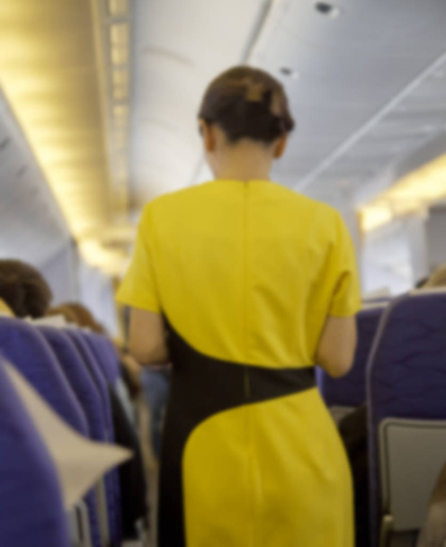wazig interieur van commerciële vliegtuig met attandant van de vlucht met passagiers op zitplaatsen tijdens de vlucht. Stewardess in donker geel uniform lopen van het gangpad. Horizontale samenstelling. - Foto, afbeelding