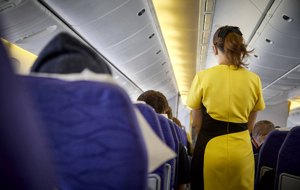 Intérieur de l'avion commercial avec passagers en service actif sur les sièges pendant le vol. Hôtesse en uniforme jaune foncé marchant dans l'allée. Composition horizontale
. - Photo, image