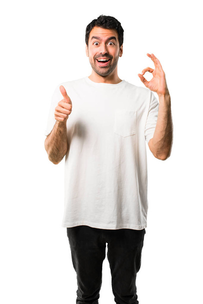 Young man with white shirt showing an ok sign with fingers and giving a thumb up gesture with the other hand on isolated white background - Zdjęcie, obraz