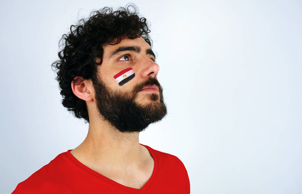 Los fanáticos del deporte se sienten orgullosos al escuchar el himno de su país. Hombre con la bandera de Egipto maquillaje en su cara y camiseta roja
.                                - Foto, Imagen