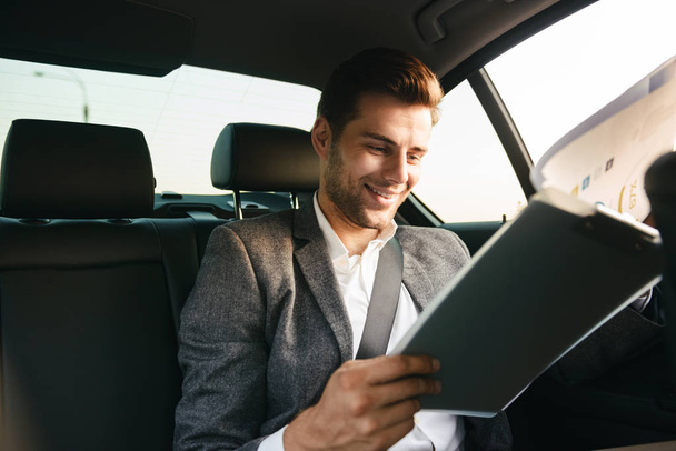 Handsome businessman looking through documents while sitting at the back of a car - Foto, Imagem