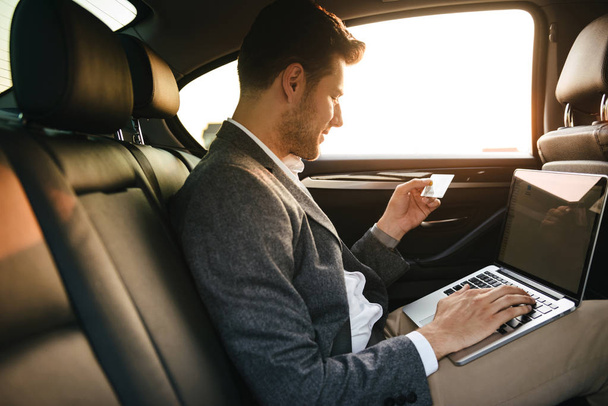 Empresário atraente usando cartão de crédito de plástico enquanto sentado com computador portátil na parte de trás de um carro
 - Foto, Imagem