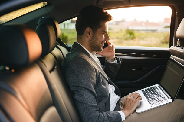 Confident businessman talking on mobile phone while sitting with laptop computer at the back of a car - Фото, изображение