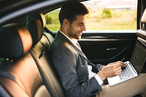Smiling businessman using plastic credit card while sitting with laptop computer at the back of a car - Фото, изображение