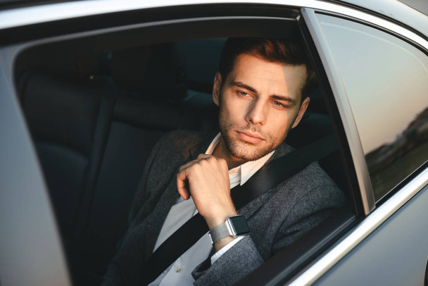 Portrait of handsome caucasian man wearing businesslike suit back sitting while riding in car with safety belt - Fotó, kép