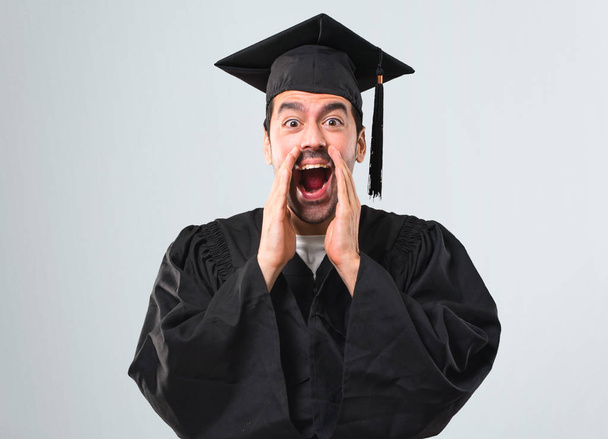 Homem em seu dia de formatura Universidade gritando com a boca bem aberta e anunciando algo sobre fundo cinza
 - Foto, Imagem