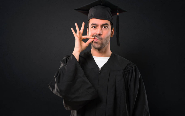 Mies hänen valmistumispäivänä University osoittaa merkki sulkemalla suun ja hiljaisuus ele tekee kuin sulkemalla suunsa vetoketju mustalla pohjalla
 - Valokuva, kuva