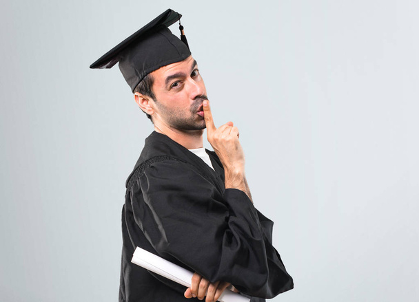 Man on his graduation day University showing a sign of closing mouth and silence gesture putting finger in mouth on grey background - Фото, зображення