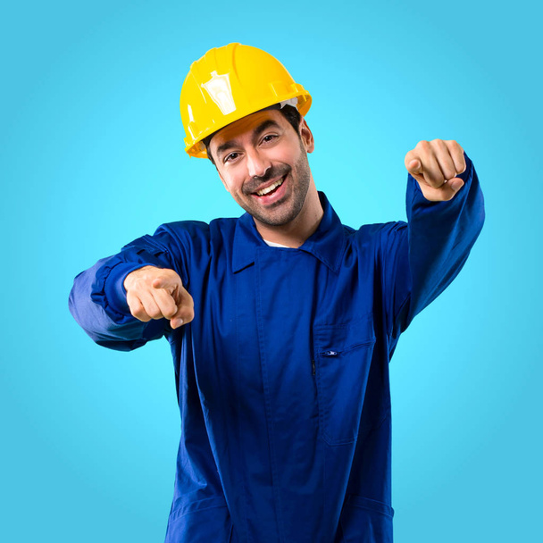 Young workman with helmet points finger at you while smiling on blue background - Фото, изображение