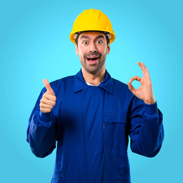 Jeune ouvrier avec casque montrant un signe ok avec les doigts et donnant un geste pouce vers le haut avec l'autre main sur fond bleu
 - Photo, image