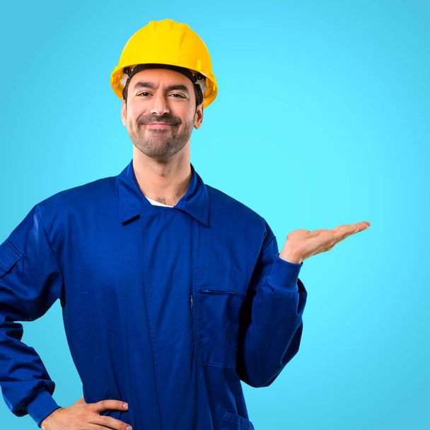 Jovem trabalhador com capacete segurando copyspace imaginário na palma da mão para inserir um anúncio no fundo azul
 - Foto, Imagem