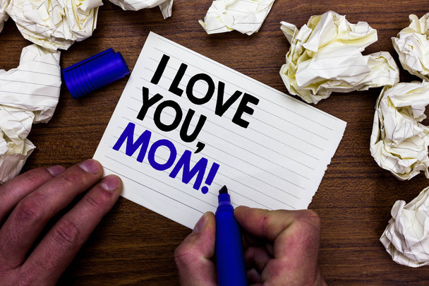 Пишу записку, показывающую "Я люблю тебя, мама". Деловое фото, демонстрирующее любовное послание, эмоциональные чувства, привязанность, теплое заявление, рукопожатие, держание маркера, написание слов, бумага, разбросанная по деревянному столу
. - Фото, изображение