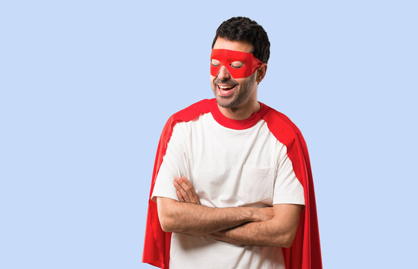Hombre superhéroe con máscara y capa roja manteniendo los brazos cruzados mientras sonríe sobre un fondo azul aislado
 - Foto, Imagen