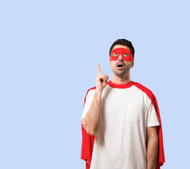 Υπερήρωα άνθρωπο με μάσκα και το κόκκινο Ακρωτήριο στέκεται και σκεφτόμαστε μια ιδέα δείχνοντας με το δάχτυλο σε απομονωμένες μπλε φόντο - Φωτογραφία, εικόνα