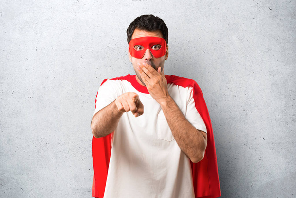 Superhero man with mask and red cape pointing with finger at someone and laughing a lot while covering mouth on textured grey background - Photo, image