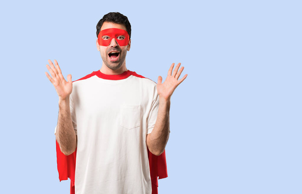Superheld man met masker en rode cape verrast en geschokt gelaatsuitdrukking. Open mond omdat niet verwachten wat er is gebeurd op geïsoleerde blauwe achtergrond - Foto, afbeelding