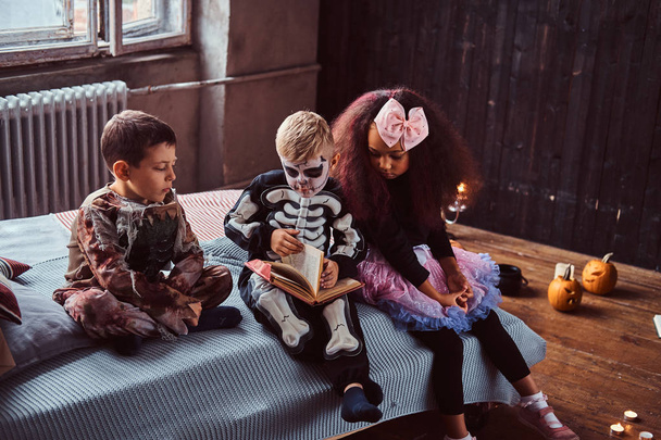 Drie multiraciale kinderen in enge kostuums horrorverhalen lezen zittend op bed in een oud huis. - Foto, afbeelding