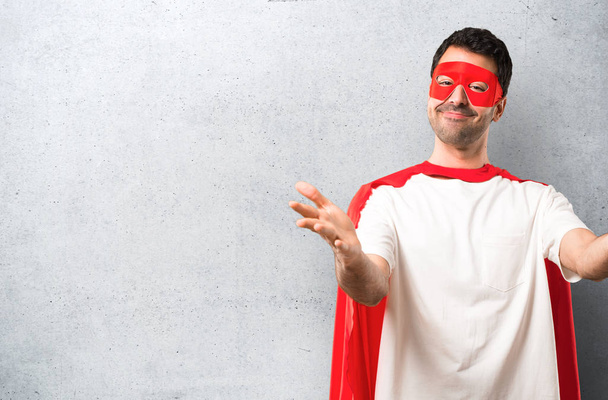 Superheld man met masker en rode cape presenteren en uitnodigen om te komen met de hand. Blij dat je kwam op gestructureerde grijze achtergrond - Foto, afbeelding