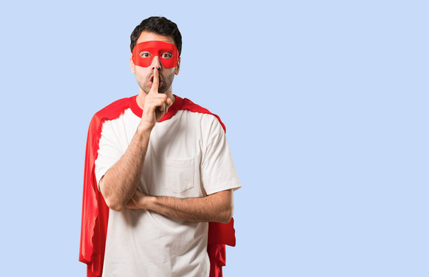 Hombre superhéroe con máscara y capa roja que muestra un signo de cerrar la boca y el gesto de silencio poniendo el dedo en la boca sobre un fondo azul aislado
 - Foto, Imagen